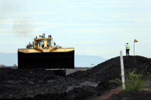 Uma escavadeira gigante trabalhando em um terreno escuro com um trabalhador ao lado.