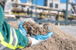 Mãos com luvas azuis segurando um material mineral em um ambiente industrial.