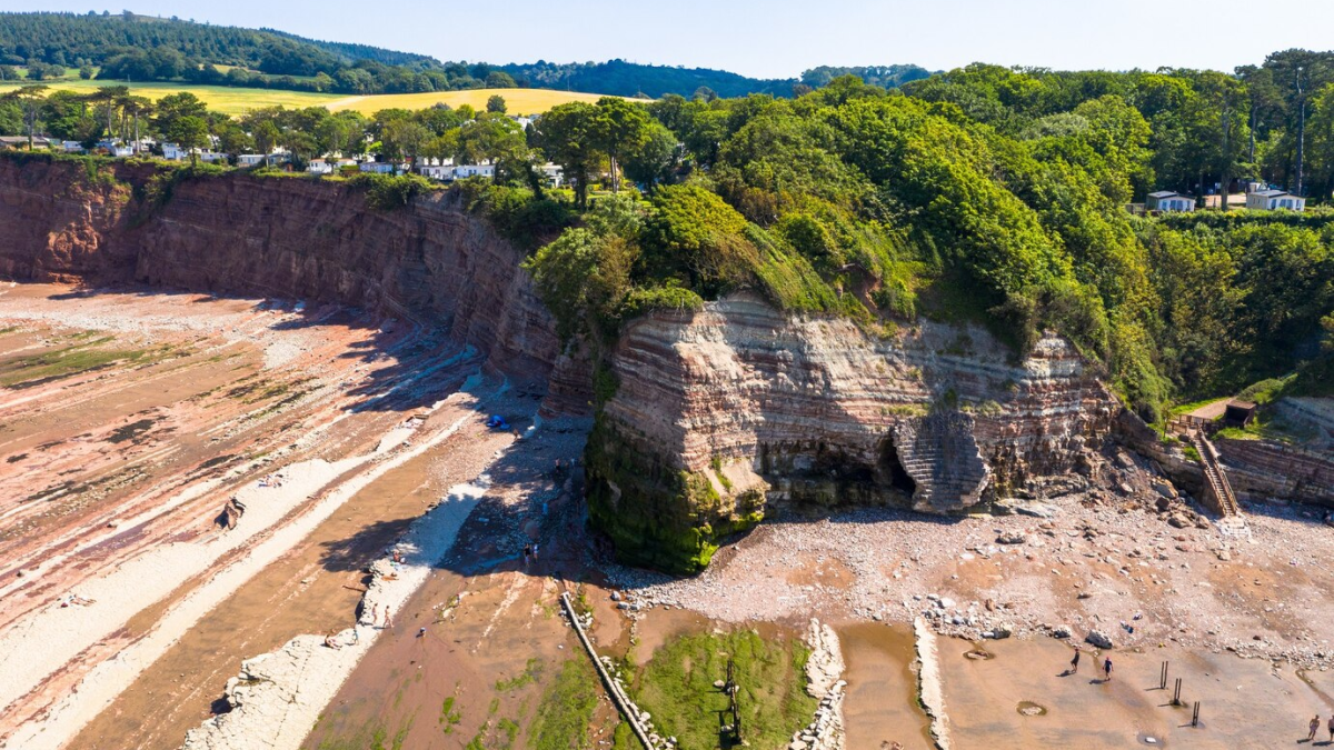 Penhascos rochosos com uma praia de areia e cascalho ao fundo, cercada por vegetação densa.