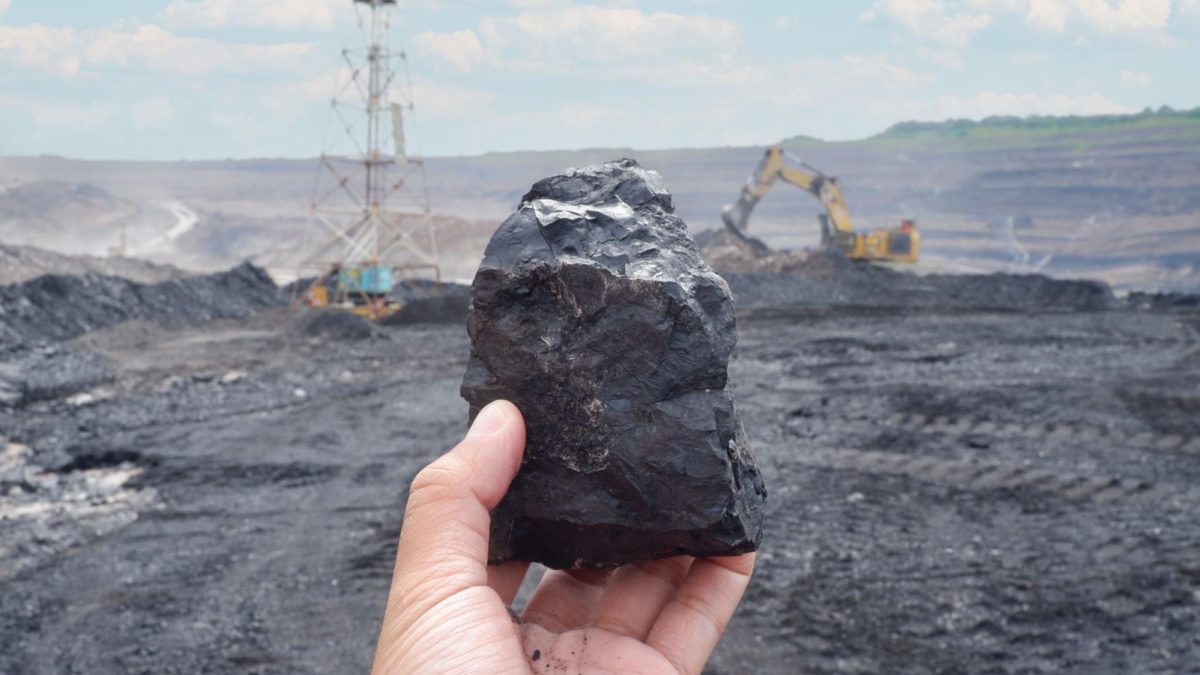 Mão segurando um pedaço de carvão em frente a uma área de mineração.