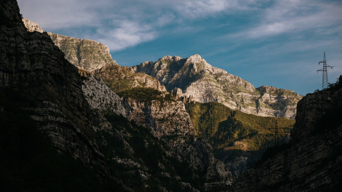 Montanhas com paredes rochosas e vegetação sob um céu azul, com uma torre de transmissão ao fundo.