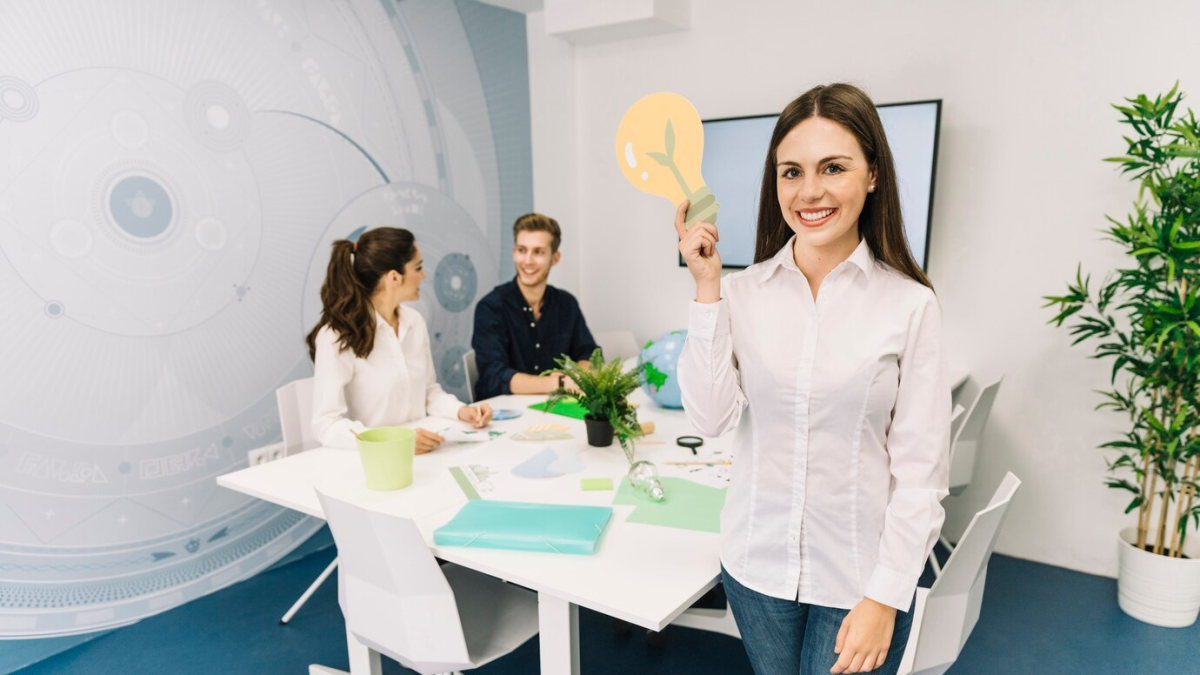 Mulher segurando uma lâmpada em forma de ideia, enquanto dois colegas conversam ao fundo.